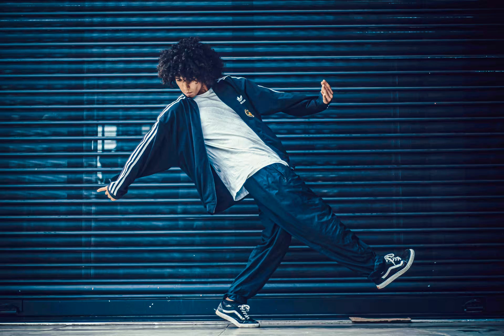 A man with an afro hairstyle is captured mid-motion in a dynamic pose, wearing a dark navy blue track jacket with white stripes, a white t-shirt, dark navy track pants, and black-and-white sneakers, set against a dark blue metal shutter background.
