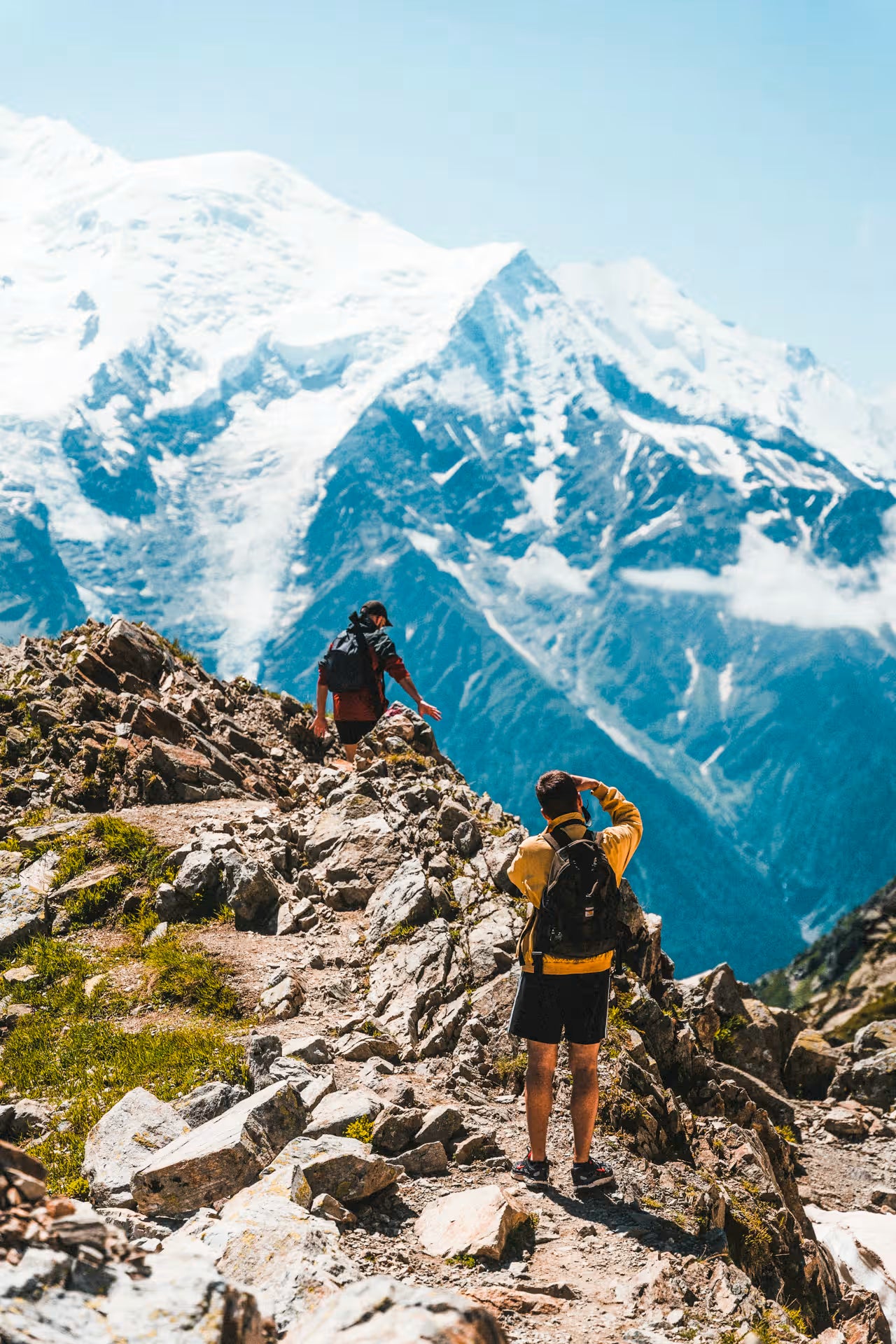 Two hikers with backpacks ascend a rocky mountain ridge with a dramatic snow-capped glacier and blue sky in the background.