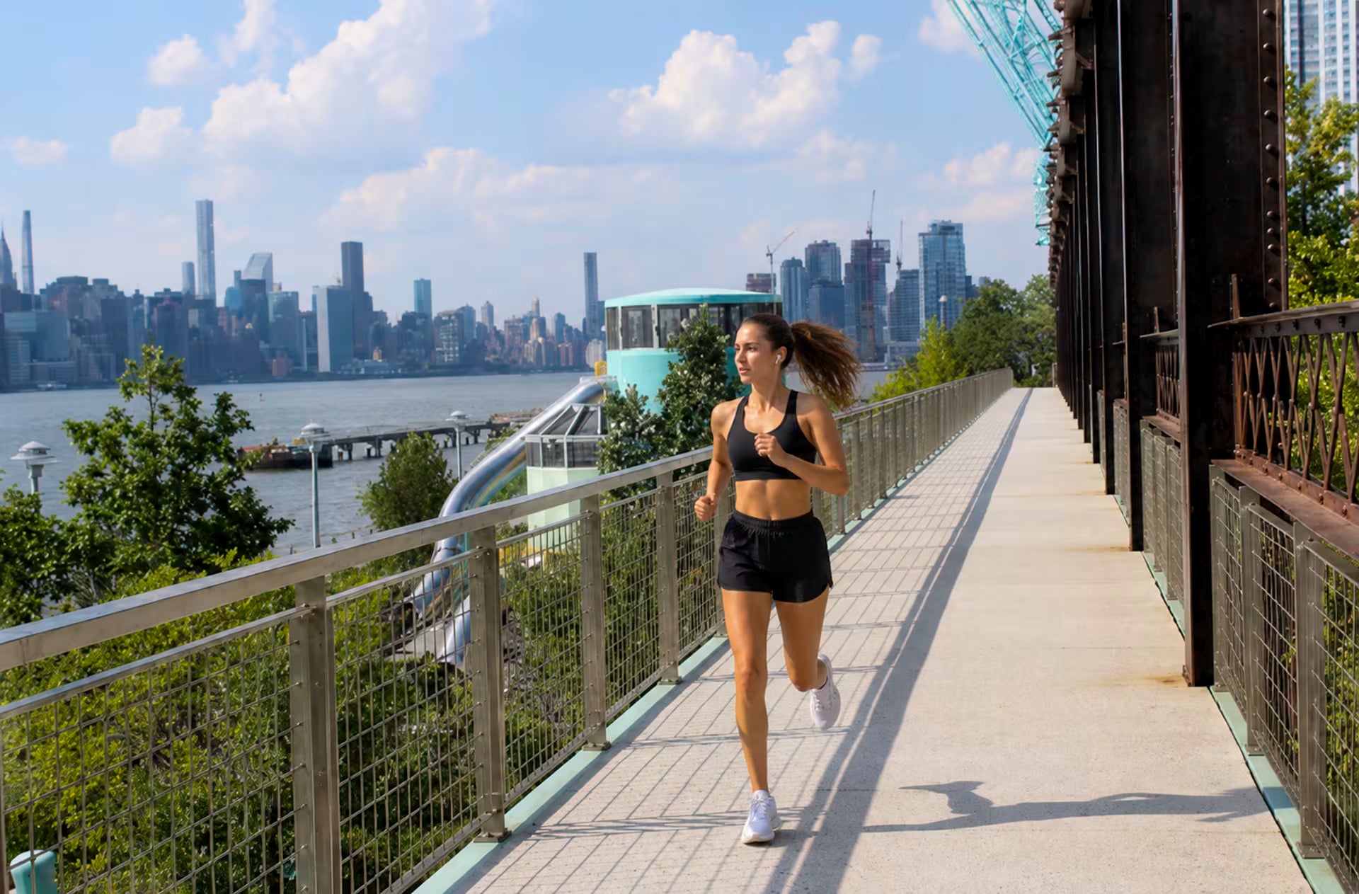 A woman in a black sports bra and black shorts is jogging on a paved path next to a waterfront city skyline, with a bridge and greenery in the background.