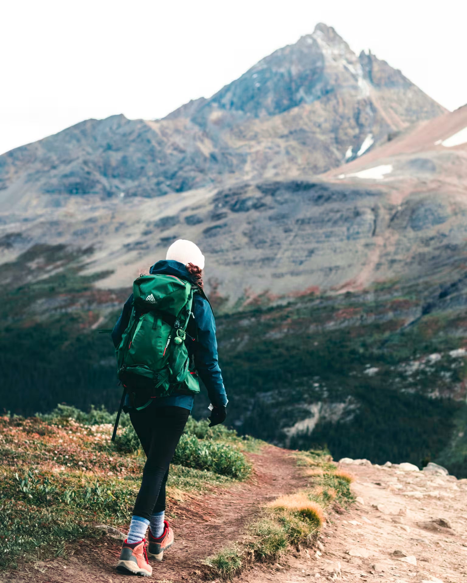 A hiker seen from behind on a mountain trail, wearing a green backpack, blue jacket, black leggings, and hiking boots, with a large rocky mountain peak in the background.