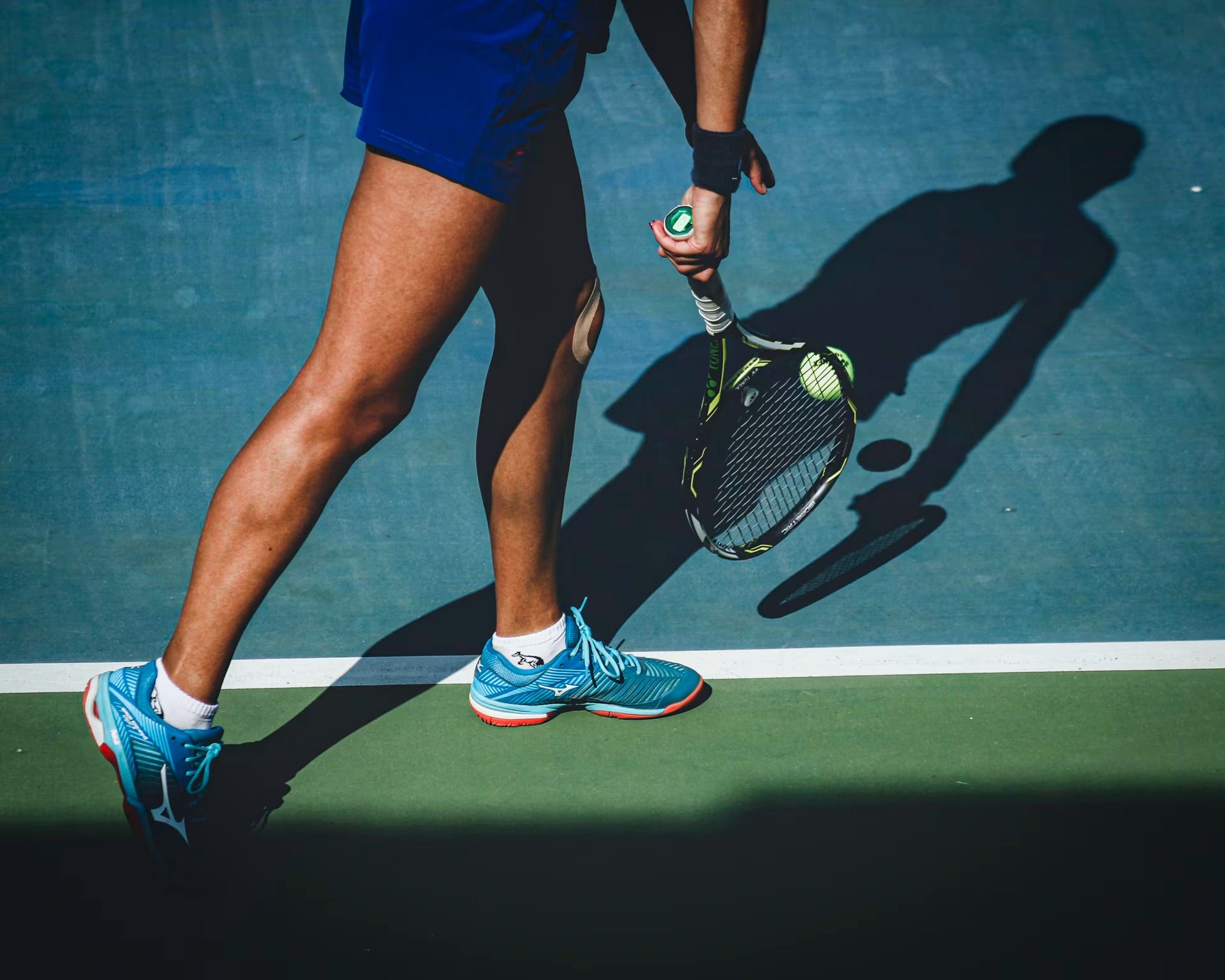 A tennis player in a blue skirt and bright blue athletic shoes holds a tennis racket and ball on a blue hard court, with strong sunlight casting a sharp shadow.