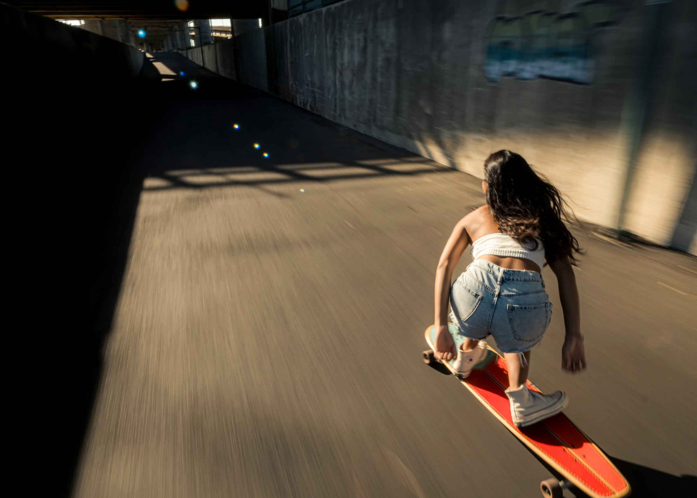 A woman rides a red skateboard down a sunlit concrete tunnel, viewed from behind. She is wearing a white crop top, light blue denim shorts, and white sneakers.