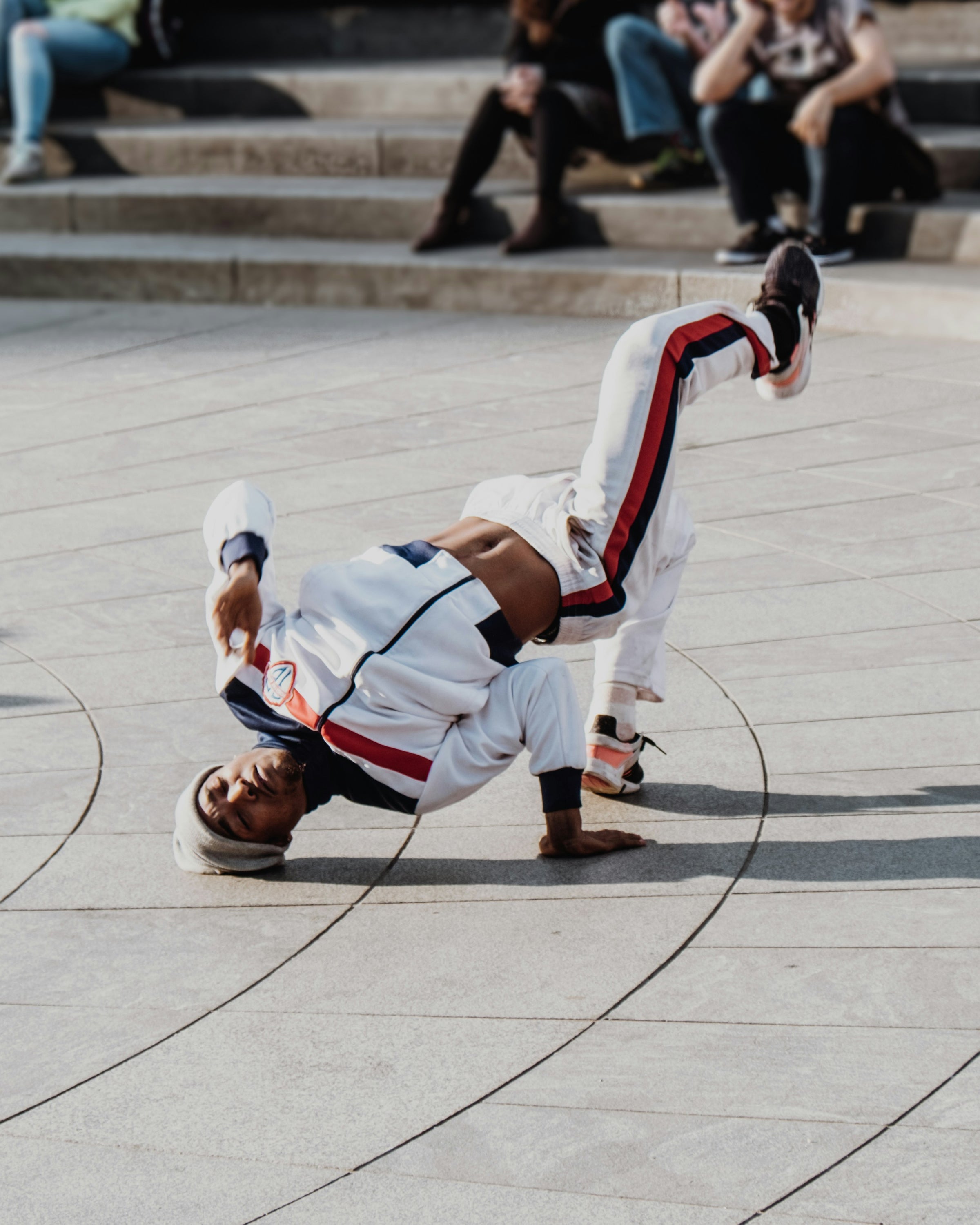 A person in a white athletic outfit with red and navy stripes performs a windmill breakdance pose on a light stone plaza, with blurred seated spectators in the background.